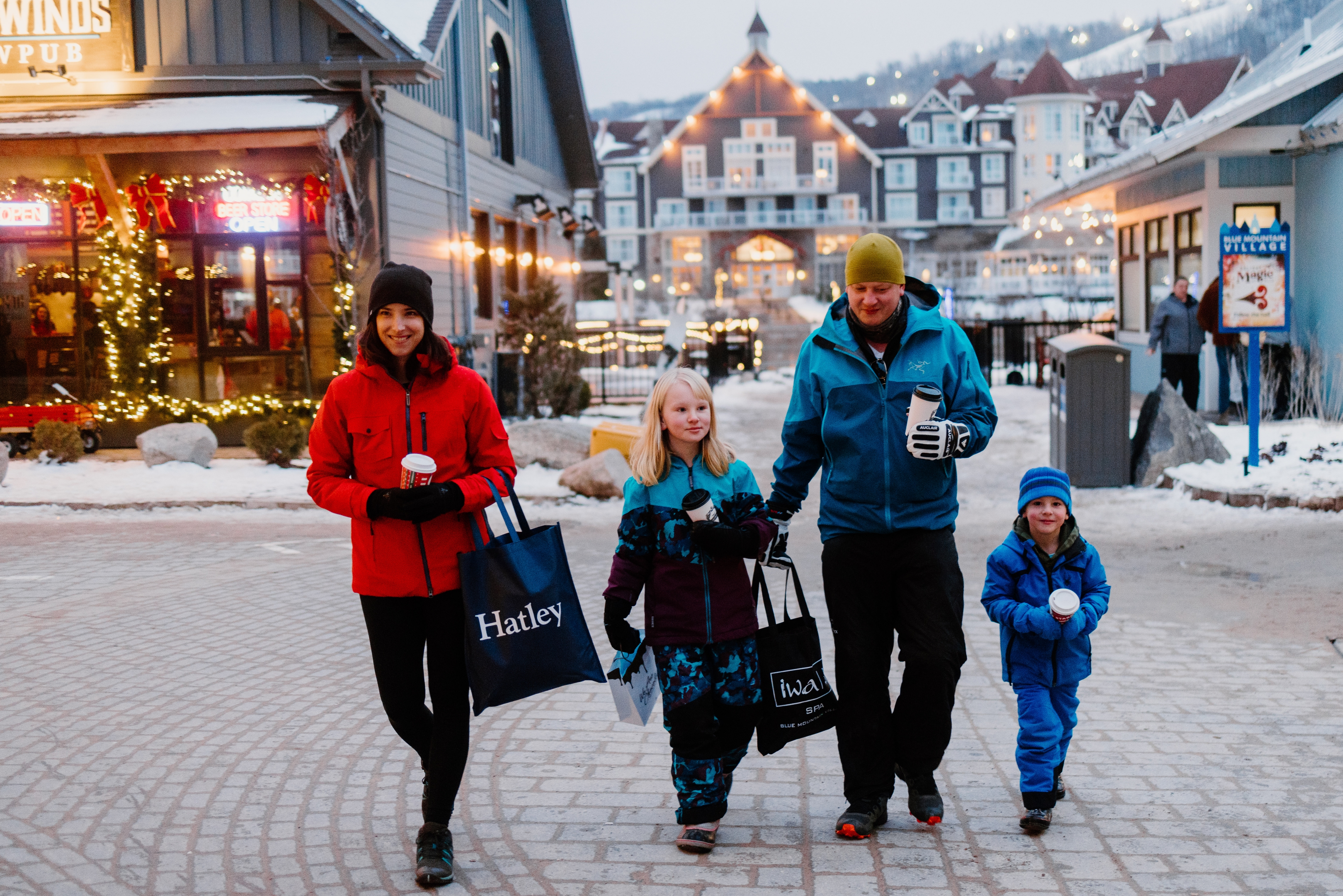 Family of four strolling through the festively lit pedestrian Village, sipping coffee and hot chocolate while shopping along the Holiday Magic Light Trail