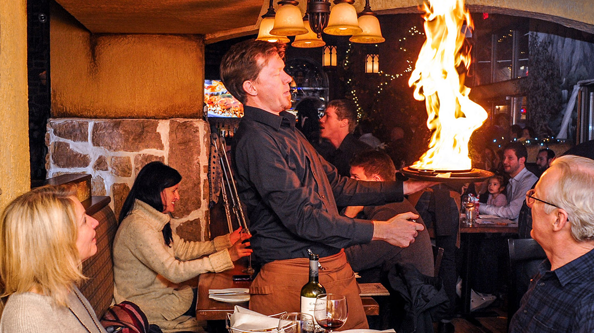 A large flame bursts as a server flambés traditional saganaki tableside at Tholos Greek Restaurant