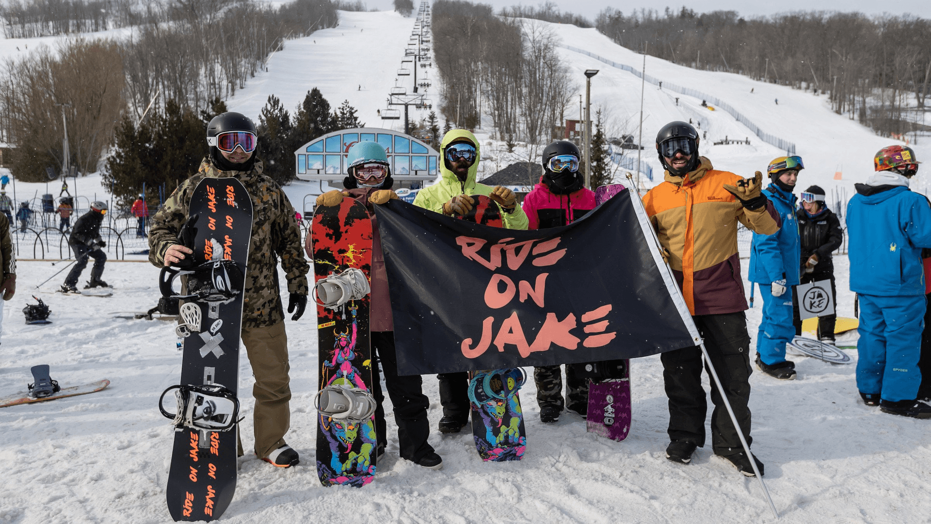 A group of snowboarders pose at the bottom of the mountain carrying "A Day For Jake" flag.