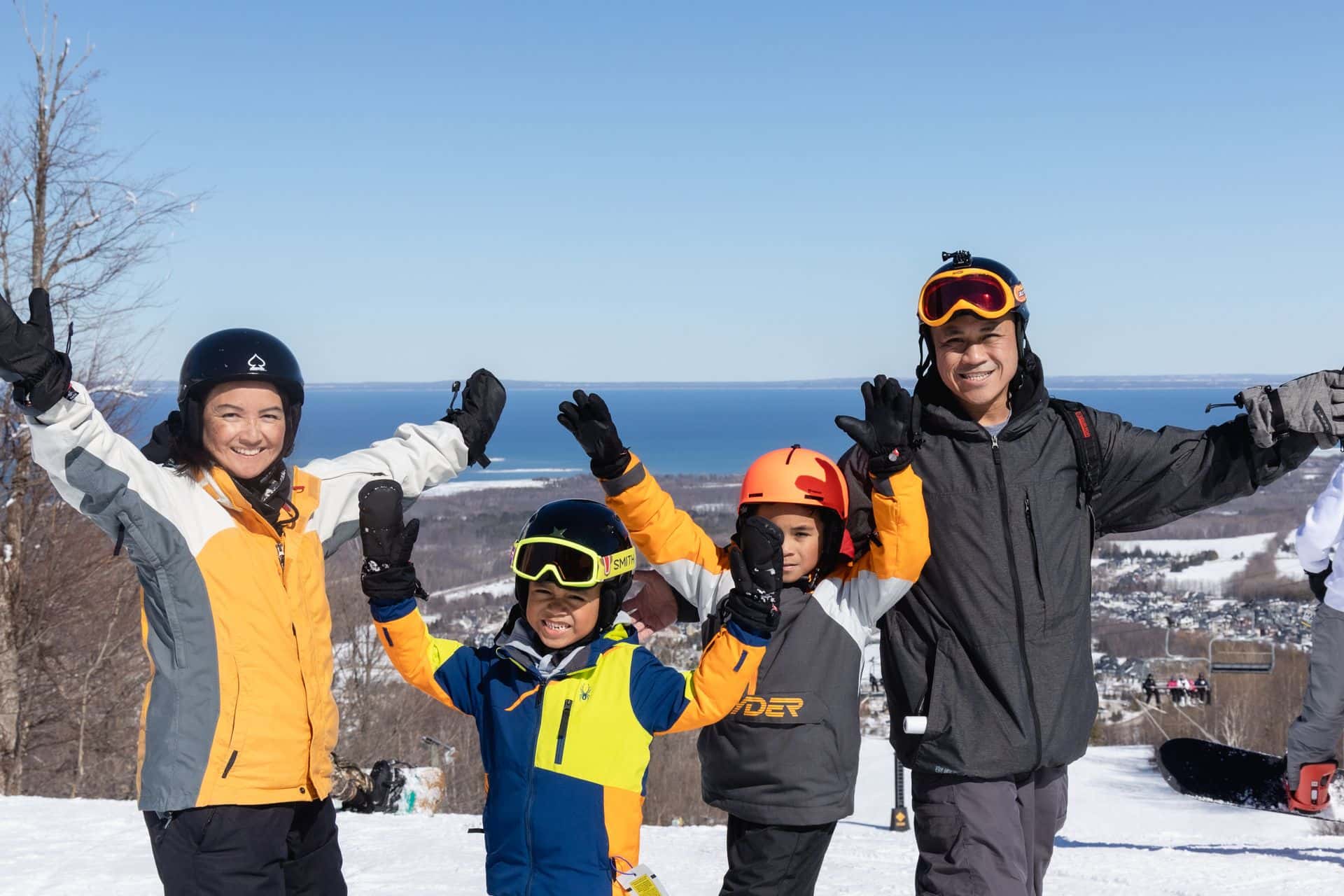 Family of four at the top of the mountain, smiling with their hands in the air, and enjoying a blue bird ski day.