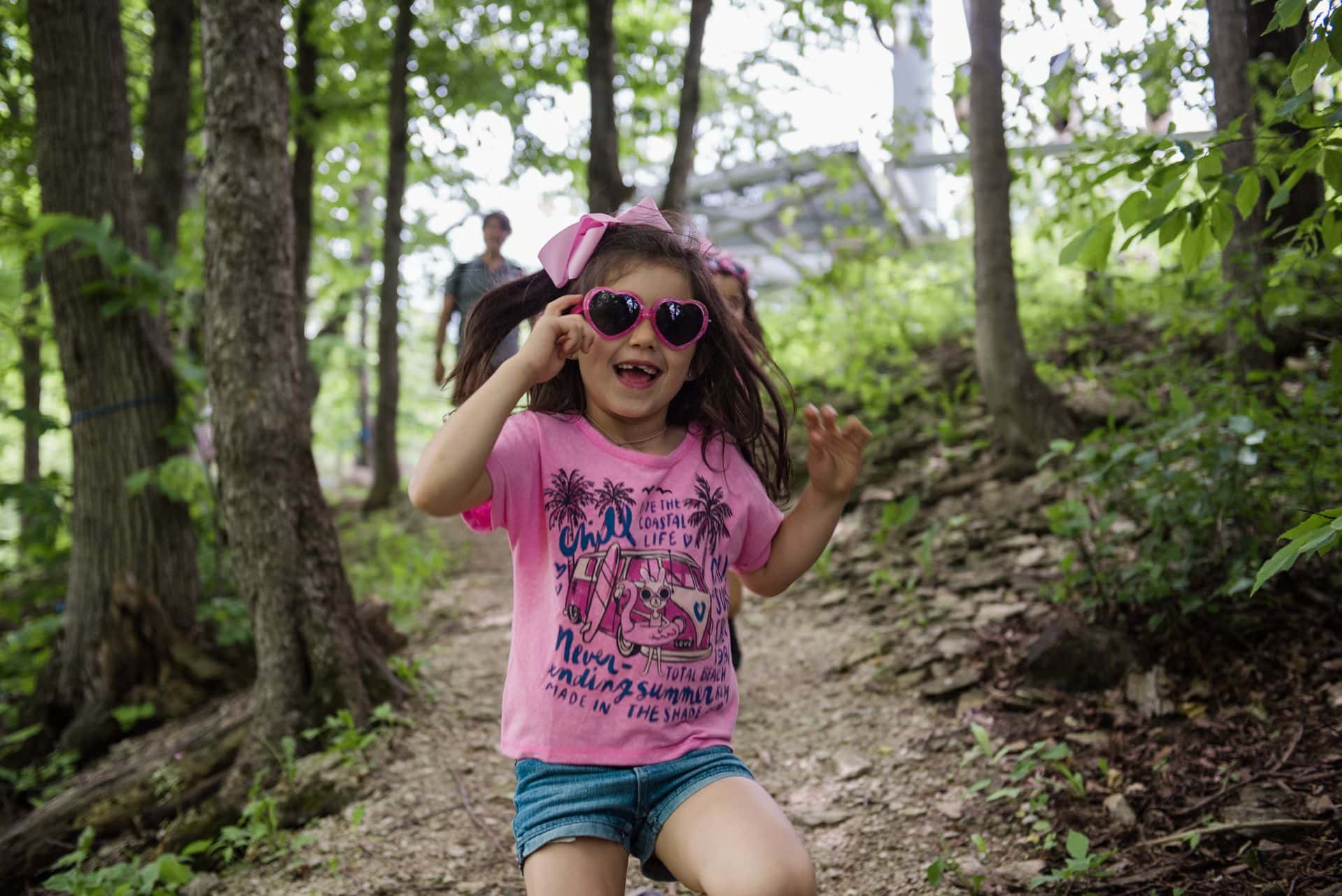 Young girl dressed in her pink outfit with matching sunglasses, laughing and smiling on a family hike in the Blue Mountain Resort Explore Park.