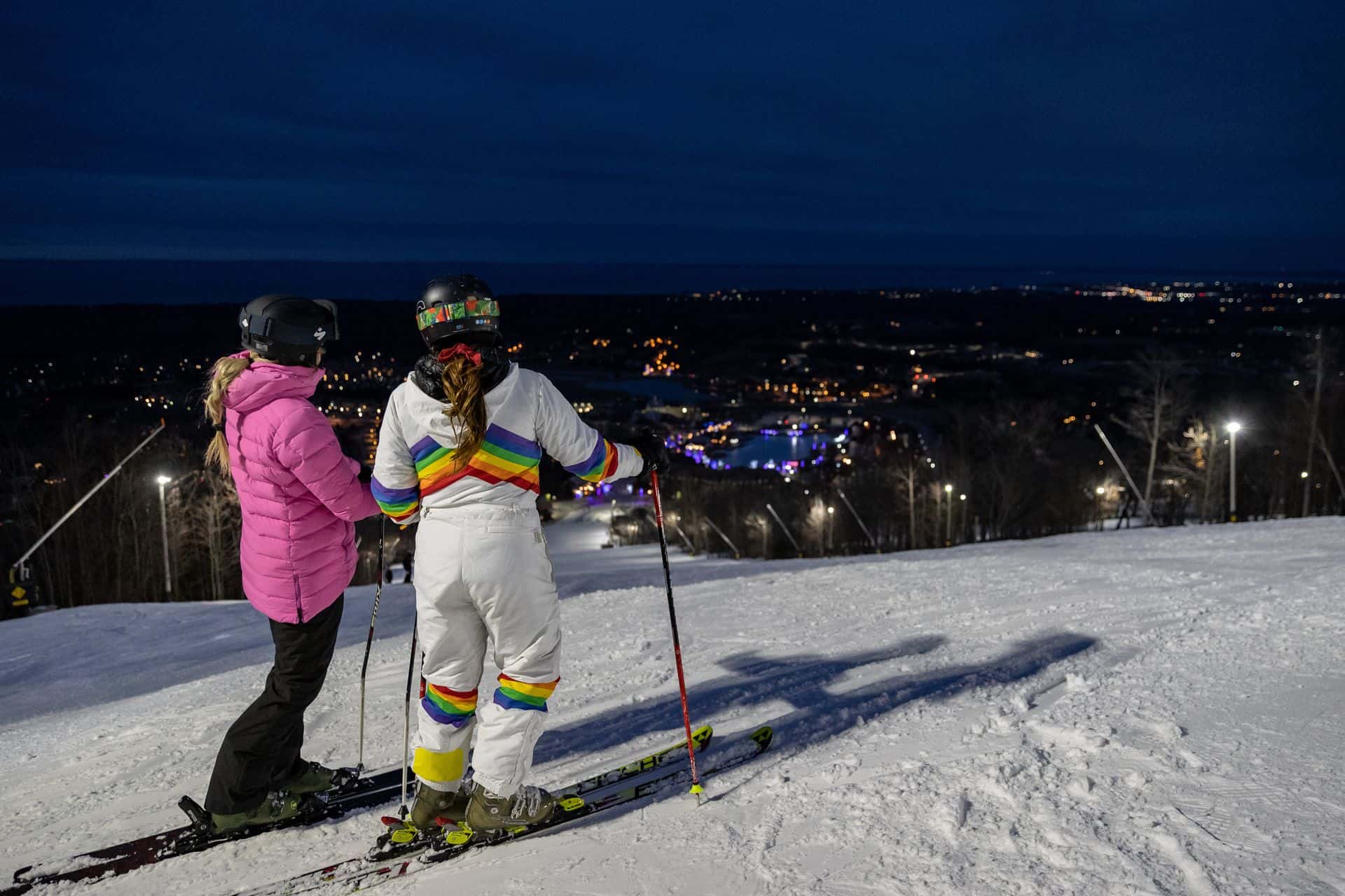 Two friends at the top of the slopes, looking out on the view of the Village lit up during night skiing. 