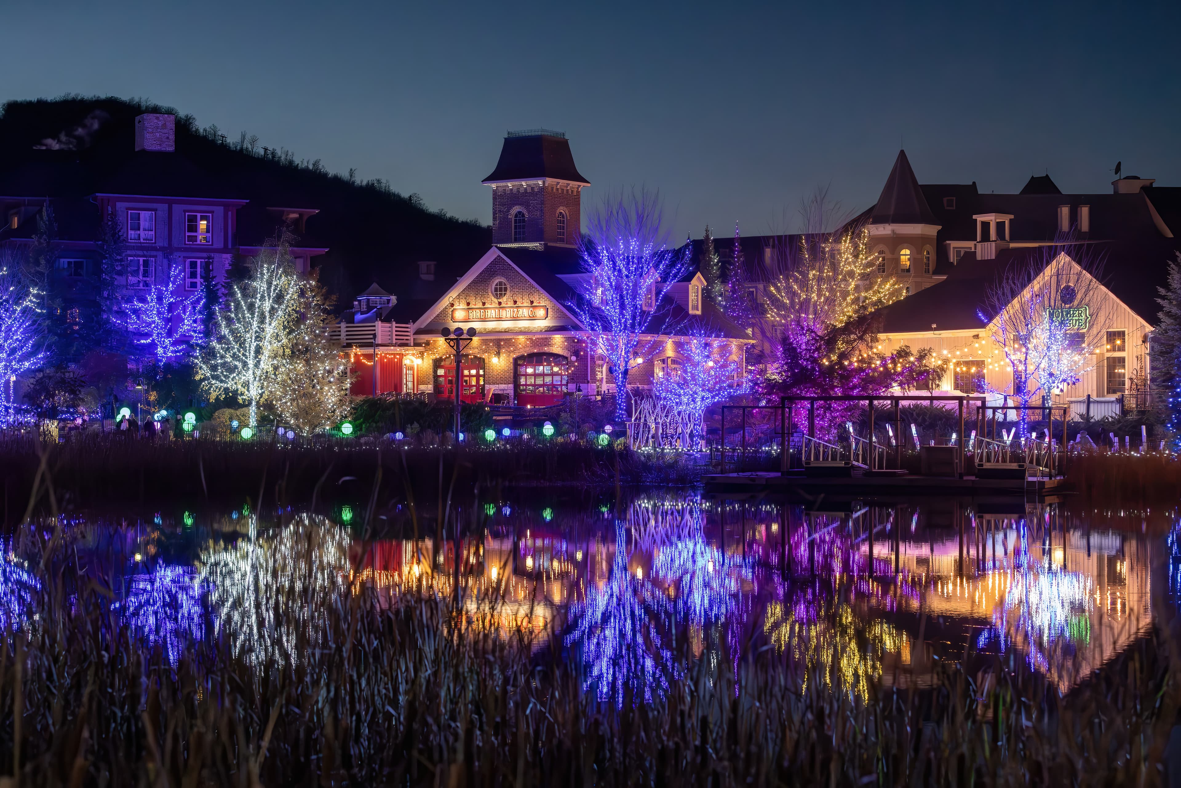 A dazzling view of the Holiday Magic Light Trail with trees and buildings decorated with lights, reflecting across the Mill Pond.
