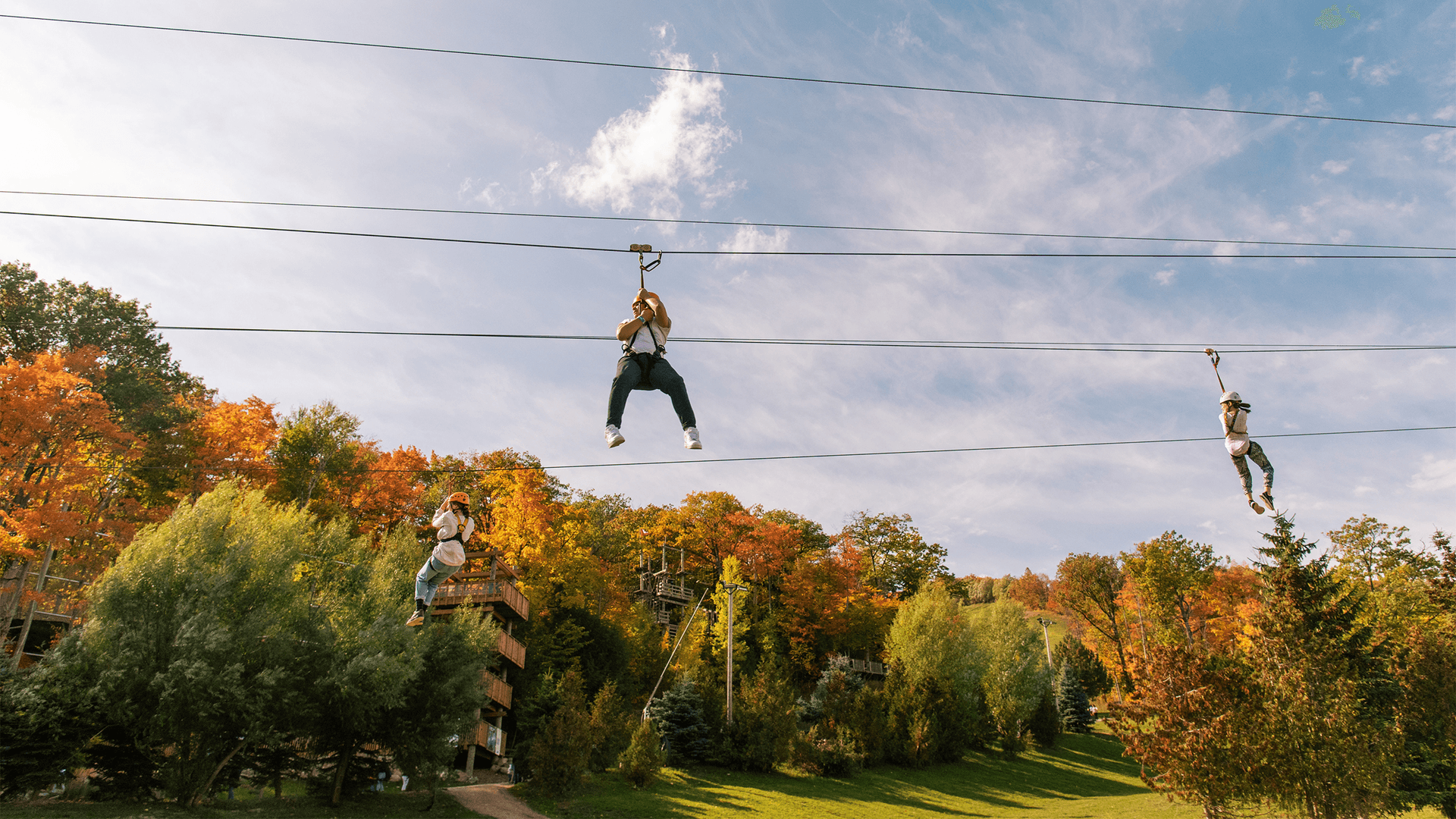 Three people glide through the amber and orange trees on the Blue Mountain Wind Rider Triple Zip course on a sunny Fall afternoon.