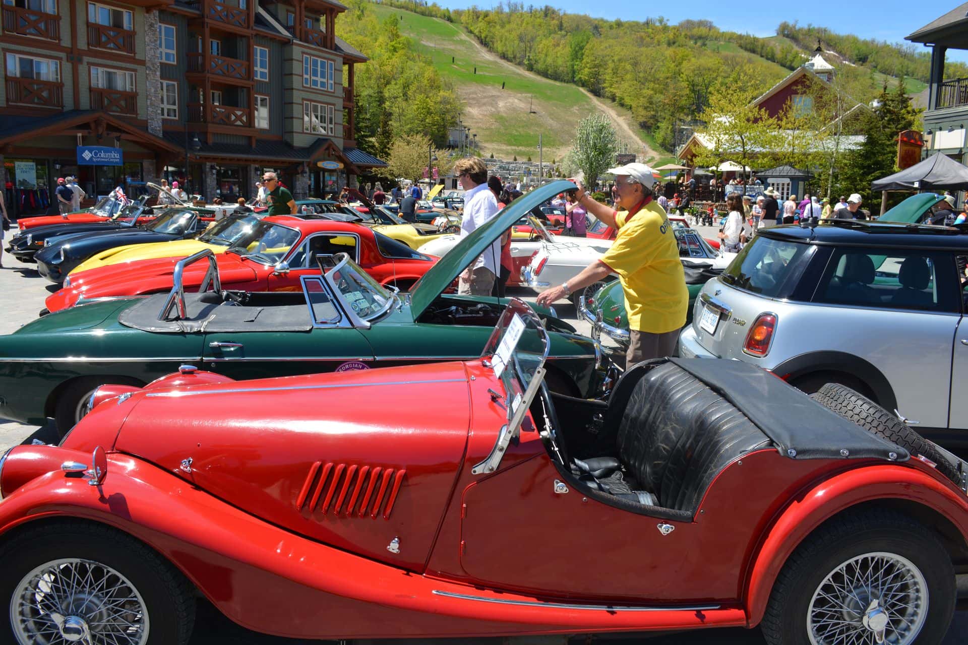 A man popping the hood of his classic car, surrounded by a village plaza full of classic cars and spectators during the British Car Show