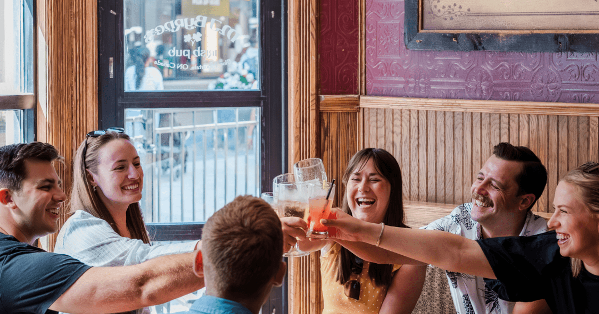 A group of six smiling friends gathered around the dining booth at MJ Byrne's Irish Pub, leaning in to clink their glasses together.