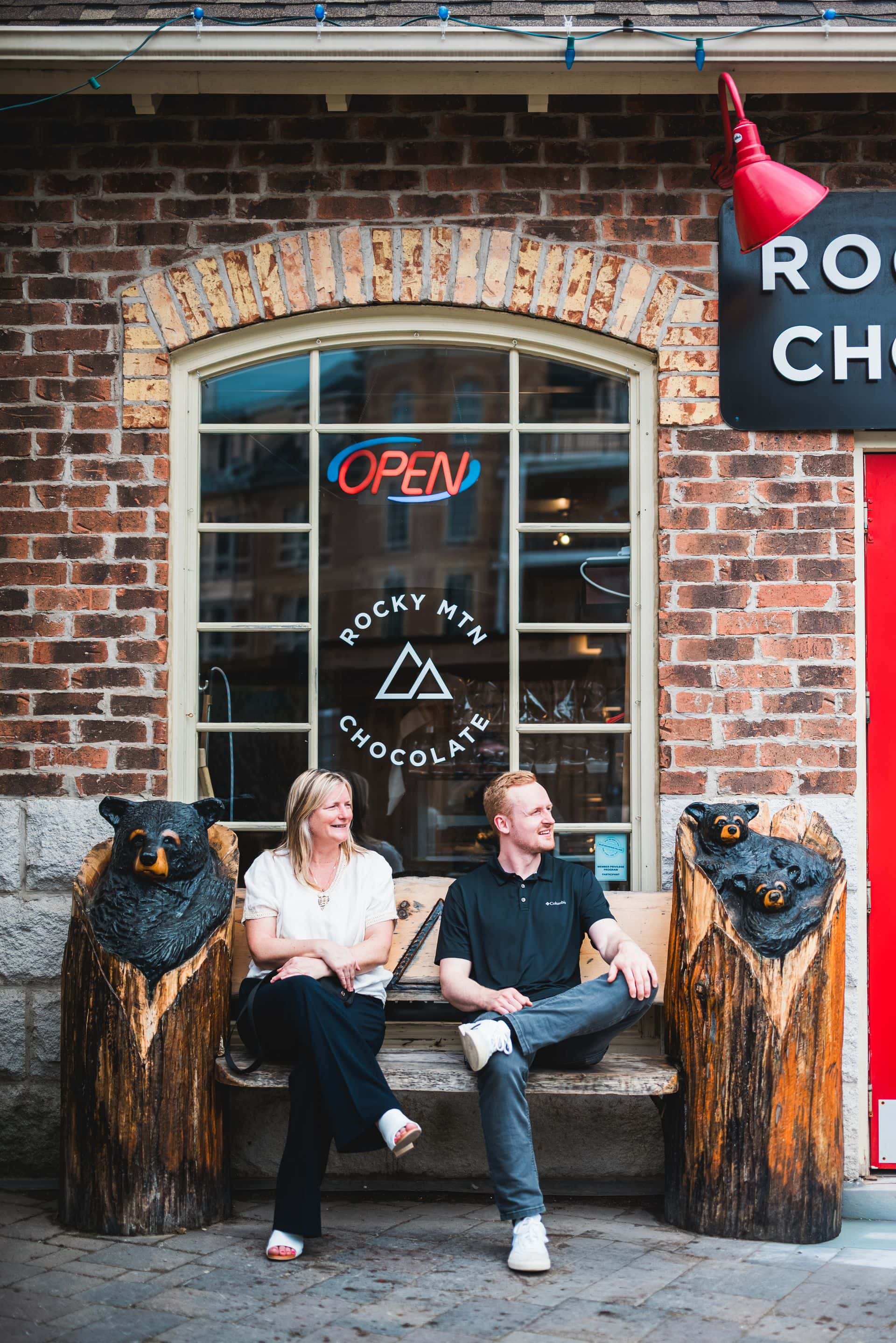 Two people sitting on the bear-carved bench outside of the Rocky Mountain Chocolate Factory storefront in Blue Mountain Village.