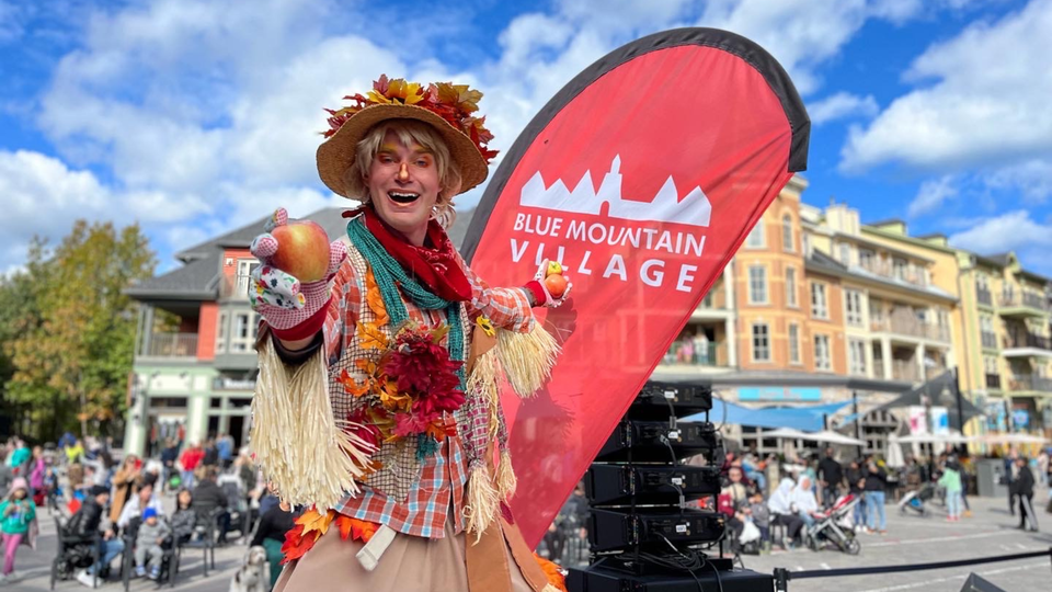 A scarecrow on stilts poses after their apple juggling routine, standing in front of a red BMVA sign on a blue sky day in the village plaza during Apple Harvest Festival Weekend at Blue.