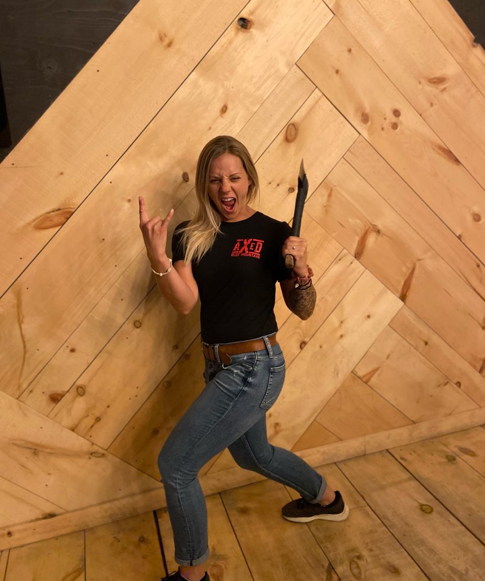 A woman standing with axe in one hand and giving rock n roll horns in the other at Axed at the Bunker