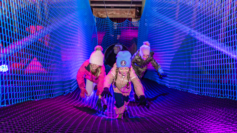 3 young girls bundled up in their winter clothes, climbing through the lit up Canopy Climb Net Adventure on a Winter's night at Blue.