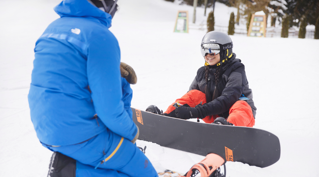 An instructor teaches a snowboarder how to do up her bindings. 