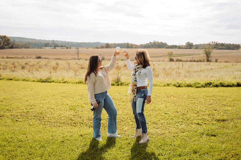 A mother and daughter clink their cider glasses together against the background of the vast farmland on a sunny fall afternoon at Windswept Cidery in Clarksburg.