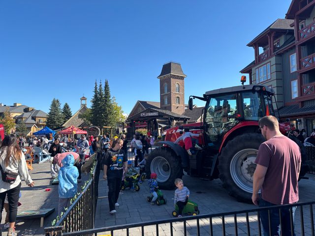 A giant industrial tractor sits in the event plaza on a sunny fall afternoon as young kids scoot around on toy tractors in the enclosed area. 