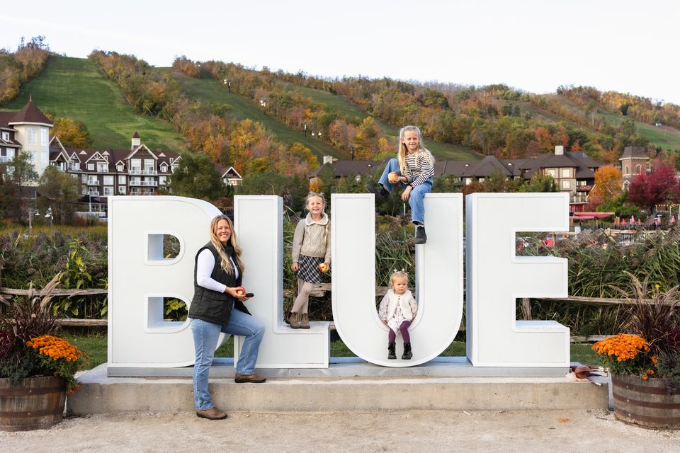A mom and her three daughters sitting and standing on different levels of the BLUE sign with views of the fall colours of the mountain in the background.