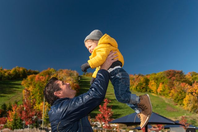A father lifts his toddler sun up in the air, with his little yellow jacket contrasting against the blue bird sky, on a Fall afternoon at Apple Harvest Festival in the Village.