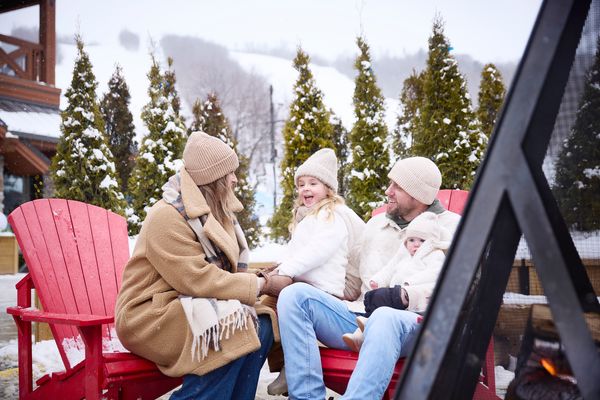 Family of four gather around an outdoor fire, sitting on red Muskoka chairs with fir trees and the snowy Mountain in the background.