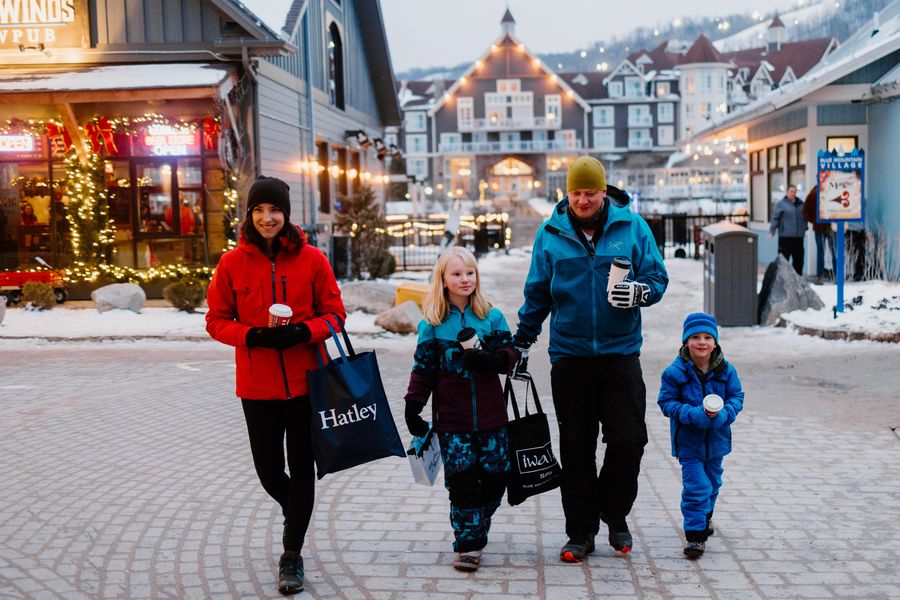 Family of four strolling through the festively lit pedestrian Village, sipping coffee and hot chocolate while shopping along the Holiday Magic Light Trail