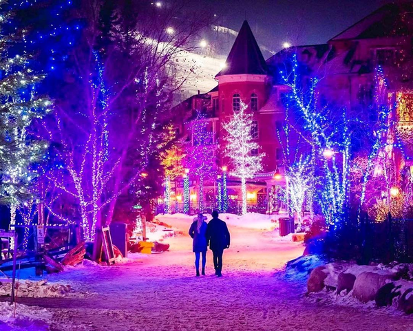 A postcard-esque photo of a couple walking along the Holiday Magic Light Trail. The snow-covered pedestrian streets are glowing in purple and blue lights