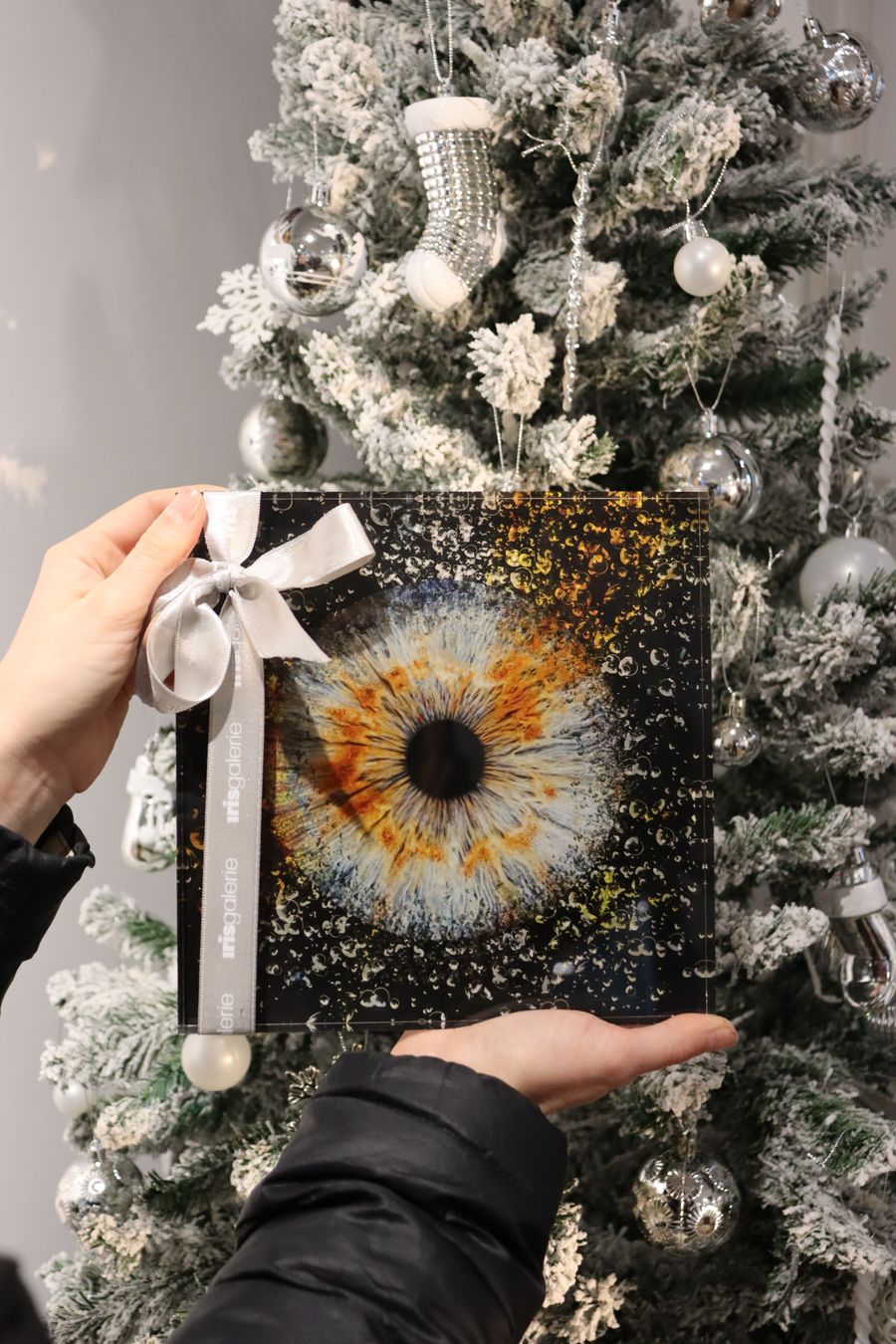 A guest in front of a decorated Christmas tree, holding up a gift wrapped photo of their iris after it was transformed into an art piece at the Iris Gallerie.