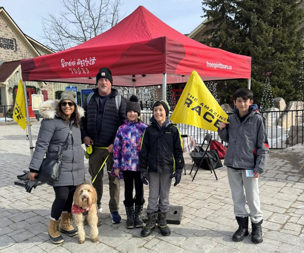 Family poses in the Village Events Plaza with The Amazing Race flag during March Break at Blue.