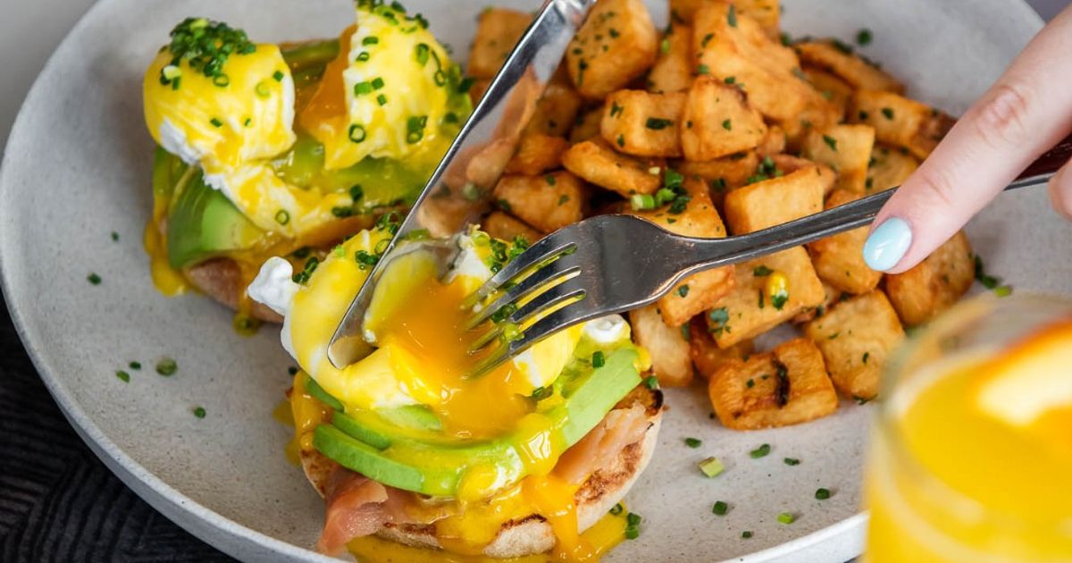 Guest breaking the yolk on their avocado eggs benedict at Oliver & Bonacini Café Grill, located in the Westin Trillium House.