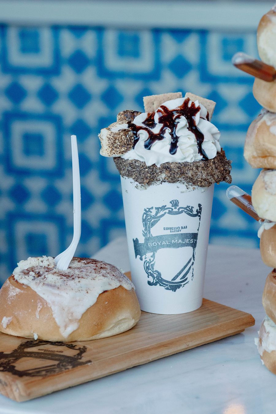 A decadent S'mores Latte from Royal Majesty, topped with whipped cream, toasted marshmallows, graham cracker, and chocolate sauce, paired with a sticky cinnamon bun.