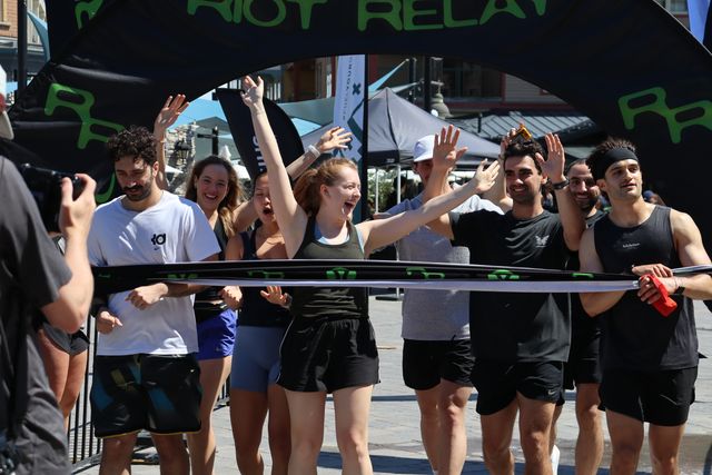 A group of racers cheer with hands in the air as they make it across the finish line after running the Riot Relay Race to Blue Mountain Village.