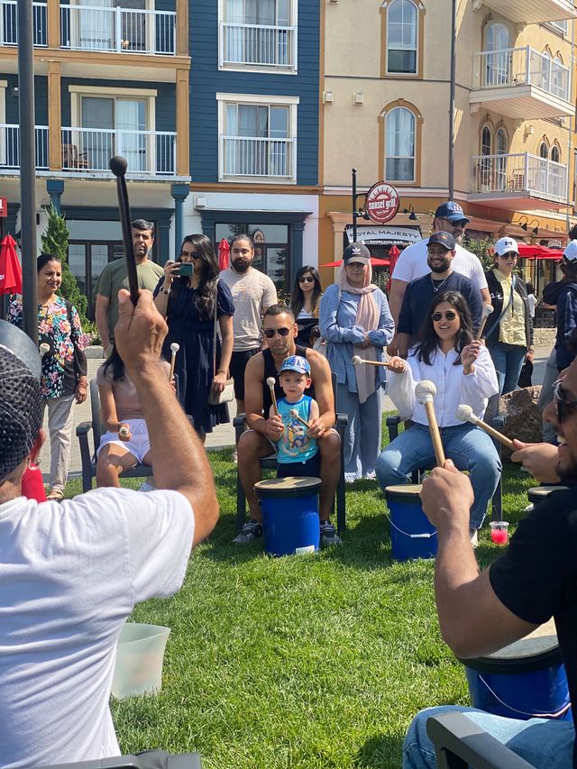 A group of people gathered in a circle, following along with Bamalamb in an interactive drumming circle on a sunny afternoon by the Village Welcome Centre.