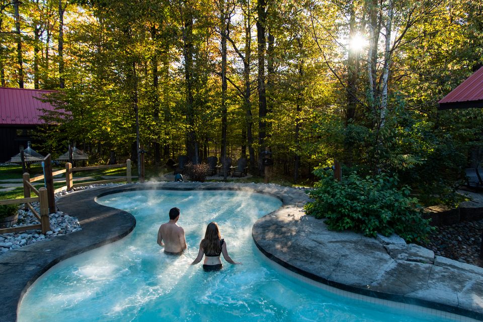 A couple walking out into the heated pool at Scandinave Spa, surrounded by a lush forest, on a sunny afternoon.