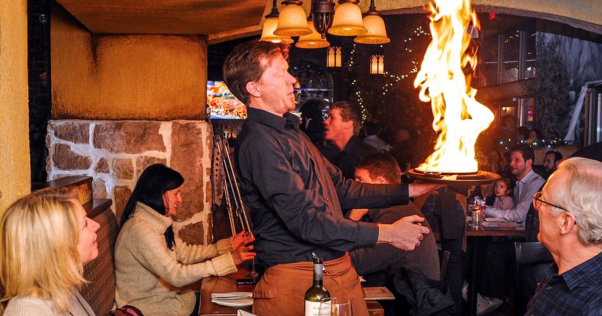 A large flame bursts as a server flambés traditional saganaki tableside at Tholos Greek Restaurant